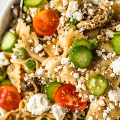 A bowl of greek pasta salad topped with sliced tomatoes, cucumbers, and feta cheese on a white background.